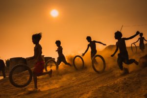 A photo of silhouettes of children rolling wheels through a dusty landscape with the sun blazing above